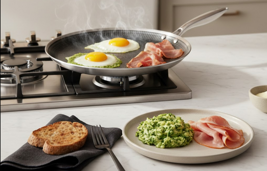 Eggs and Bacon in a griddle pan on stovetop. Foreground: plate with pesto green eggs and ham and black napkin with slice of toast and a fork